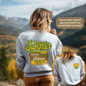 May include: A light gray sweatshirt with a green and yellow "Johnson's Summer Camp" design on the back. The design includes the text "Welcome to our adventure", "Rebecca Counselor", and "EST. 2025". A smaller badge is on the front.