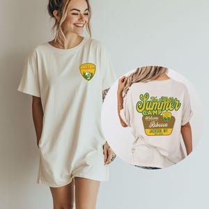 Puede incluir: Camiseta color crema con un dise&ntilde;o en el bolsillo delantero y un dise&ntilde;o en la espalda. El dise&ntilde;o frontal es una insignia de consejero amarilla y verde. El dise&ntilde;o trasero dice "The Misfit's Summer Camp" con el nombre Rebecca y la ubicaci&oacute;n Jackson, WY.