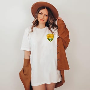 Puede incluir: Camiseta blanca con un parche amarillo y verde que dice "Camp Director". La camiseta se combina con un sombrero marr&oacute;n y una chaqueta marr&oacute;n. La camiseta es de estilo casual.