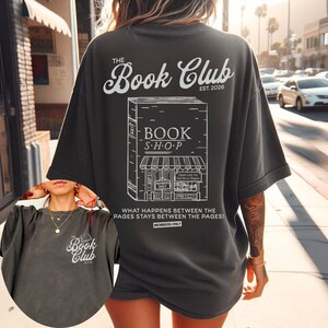 K&ouml;nnte beinhalten: Dunkelgraues T-Shirt mit wei&szlig;er Grafik eines Buchladens und dem Text "The Book Club EST. 2026." Das Shirt zeigt auch den Text "Was zwischen den Seiten passiert, bleibt zwischen den Seiten!"