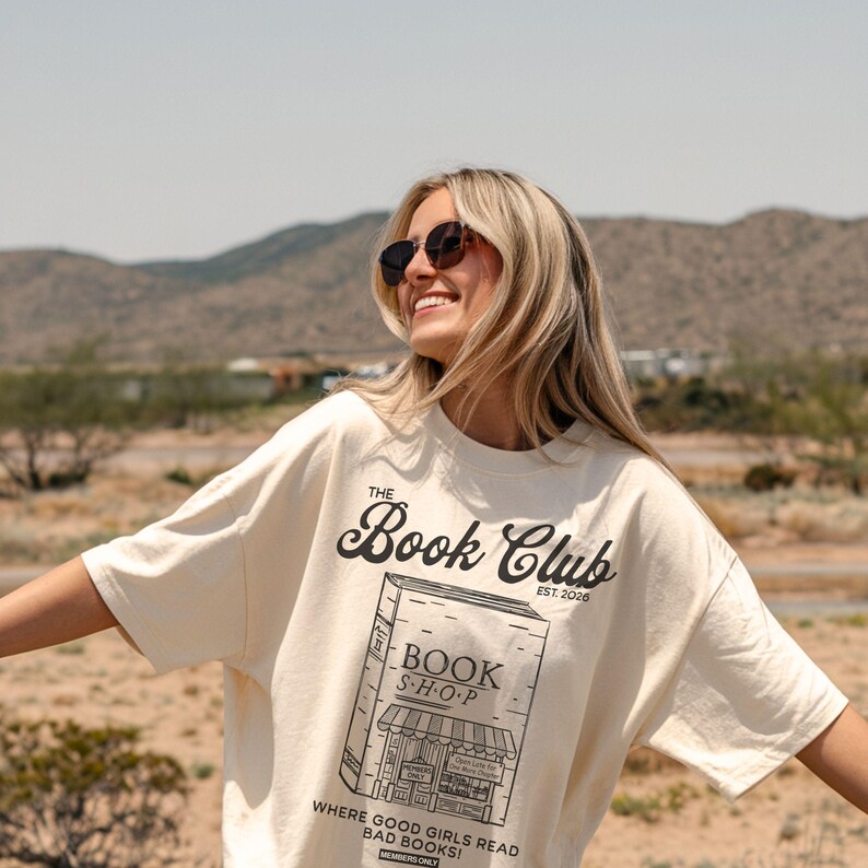K&ouml;nnte beinhalten: Cremefarbenes T-Shirt mit dem Text "The Book Club" und einer Grafik eines Buchladens. Das Shirt hat auch den Text "Where good girls read bad books!" und "Members Only". Das Shirt ist ein l&auml;ssiger Stil.