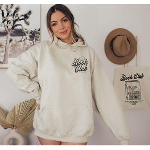 K&ouml;nnte beinhalten: Cremefarbener Kapuzenpullover mit Fronttasche und Kordelzug. Der Text "The Book Club" ist auf der Vorderseite aufgedruckt. Eine Tragetasche mit demselben Text und einer Buchladen-Illustration ist ebenfalls zu sehen. Ein brauner Hut h&auml;ngt im Hintergrund.