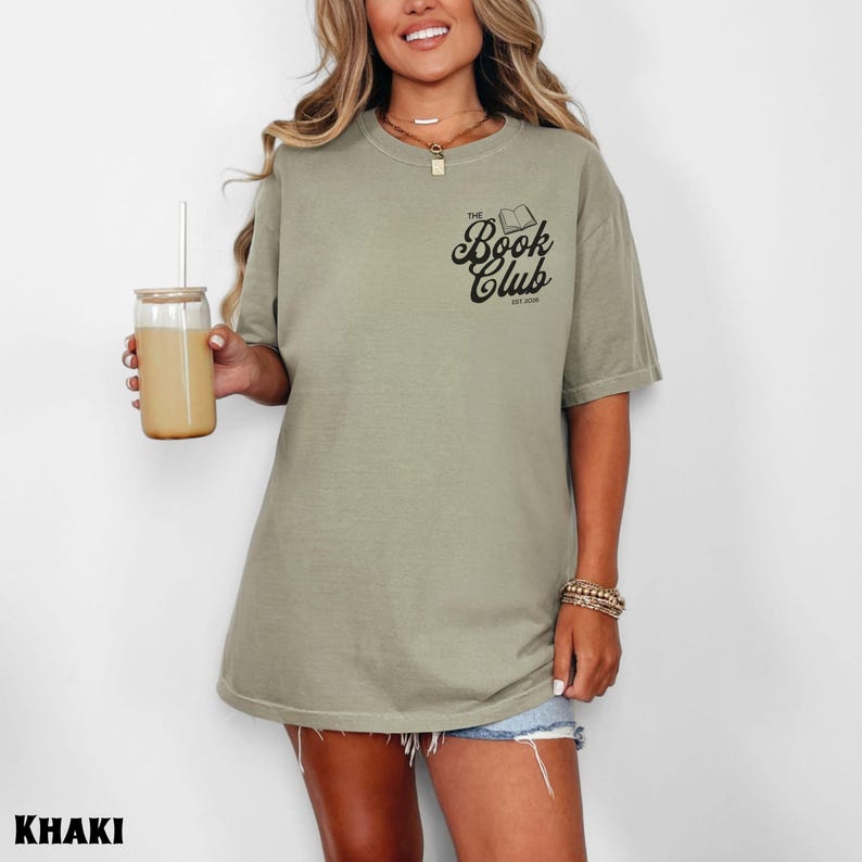 K&ouml;nnte beinhalten: Khaki-farbenes T-Shirt mit dem Text "The Book Club" und einer Buchillustration. Das Shirt hat eine lockere Passform und kurze &Auml;rmel. Ein Glas mit Strohhalm ist ebenfalls zu sehen.