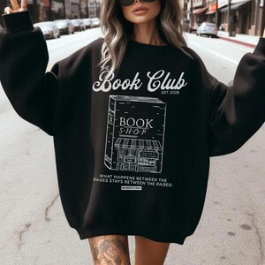 K&ouml;nnte beinhalten: Schwarzes Sweatshirt mit wei&szlig;em Text und Grafik. Der Text lautet "The Book Club EST. 2026" &uuml;ber einer Buchladen-Grafik. Darunter steht "WHAT HAPPENS BETWEEN THE PAGES STAYS BETWEEN THE PAGES!"