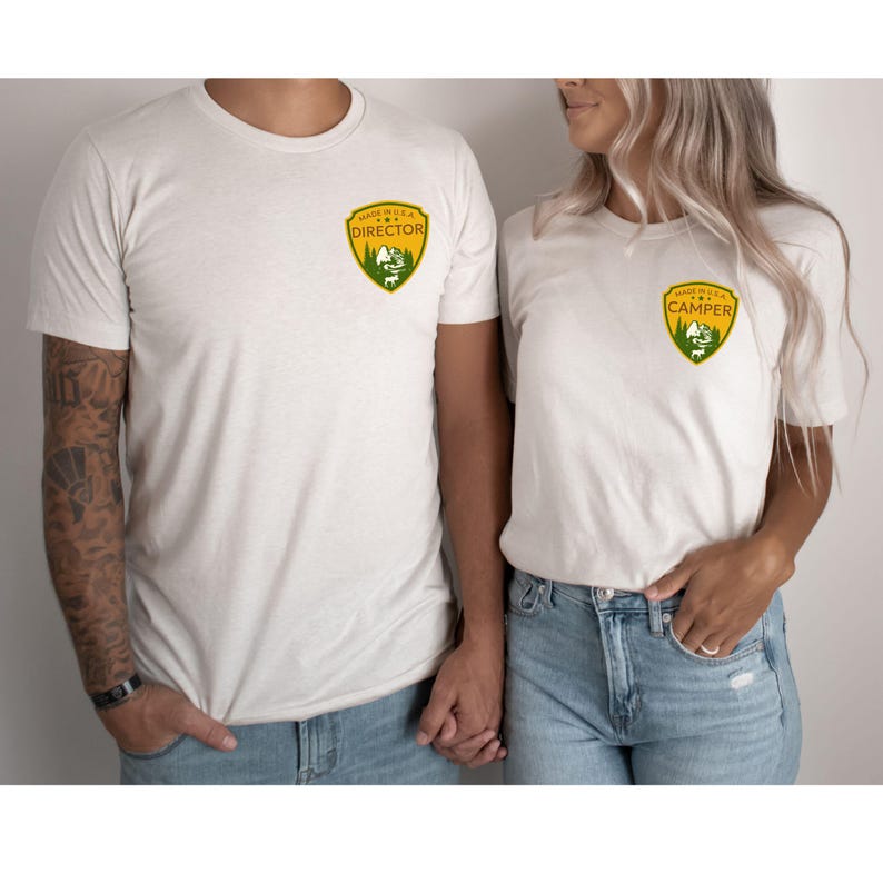 Puede incluir: Dos camisetas de color beige claro con un parche que dice "MADE IN U.S.A. DIRECTOR" y "MADE IN U.S.A. CAMPER" en un dise&ntilde;o amarillo, verde y blanco. Las camisetas las llevan dos personas, tomadas de la mano.
