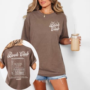 K&ouml;nnte beinhalten: Braunes T-Shirt mit dem Text "The Book Club" und einer Buchgrafik auf der Vorderseite. Auf der R&uuml;ckseite des Shirts befindet sich eine Buchladen-Grafik und der Text "What happens between the pages stays between the pages!"
