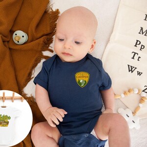 May include: A navy blue baby onesie with a "Made in the USA Camper" patch. The onesie is on a white surface with a brown blanket and a pacifier. A small white onesie with the text "Summer's Garden" is hanging on a wooden rack.