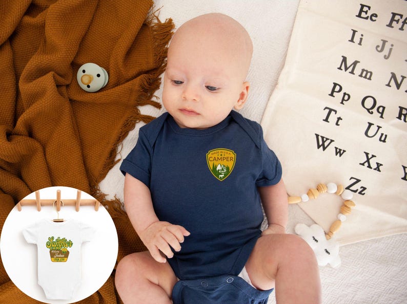 May include: A navy blue baby onesie with a "Made in the USA Camper" patch. The onesie is on a white surface with a brown blanket and a pacifier. A small white onesie with the text "Summer's Garden" is hanging on a wooden rack.