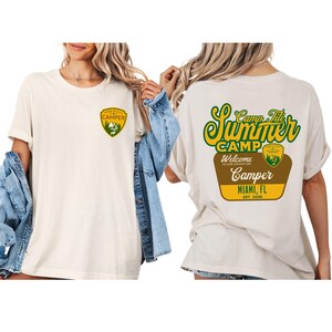 Puede incluir: Camiseta color crema con dise&ntilde;o frontal y trasero. La parte delantera presenta una insignia de campista verde y amarilla. La parte trasera muestra "Camp Tit Summer Camp" en verde y amarillo, con "Camper Miami, FL" debajo.
