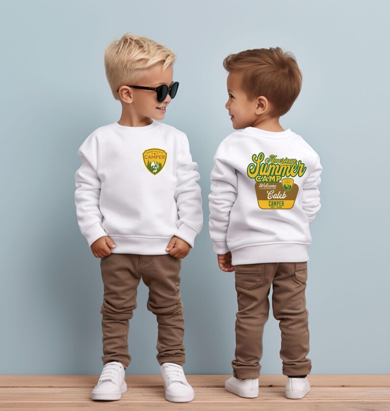 May include: Two young children wearing white sweatshirts and brown pants. The front of the sweatshirt has a green and yellow patch that says "Camper". The back of the sweatshirt has the text "American Summer Camp" and "Welcome Camper".