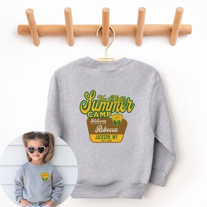 May include: A gray sweatshirt with the text "The Misfits Summer Camp" in green and yellow, with a brown and yellow badge that says "Welcome to our adventure" and "Rebecca Jackson, WY Est. 2025". The sweatshirt is hanging on a wooden rack.