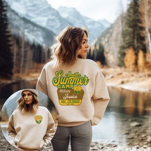 Puede incluir: Sudadera beige con un dise&ntilde;o verde y amarillo de "Smith's Summer Camp" en la espalda, incluyendo el nombre "Jessica" y "Vail, Colorado". Una versi&oacute;n m&aacute;s peque&ntilde;a del dise&ntilde;o est&aacute; en la parte delantera. El fondo muestra una monta&ntilde;a y un lago.