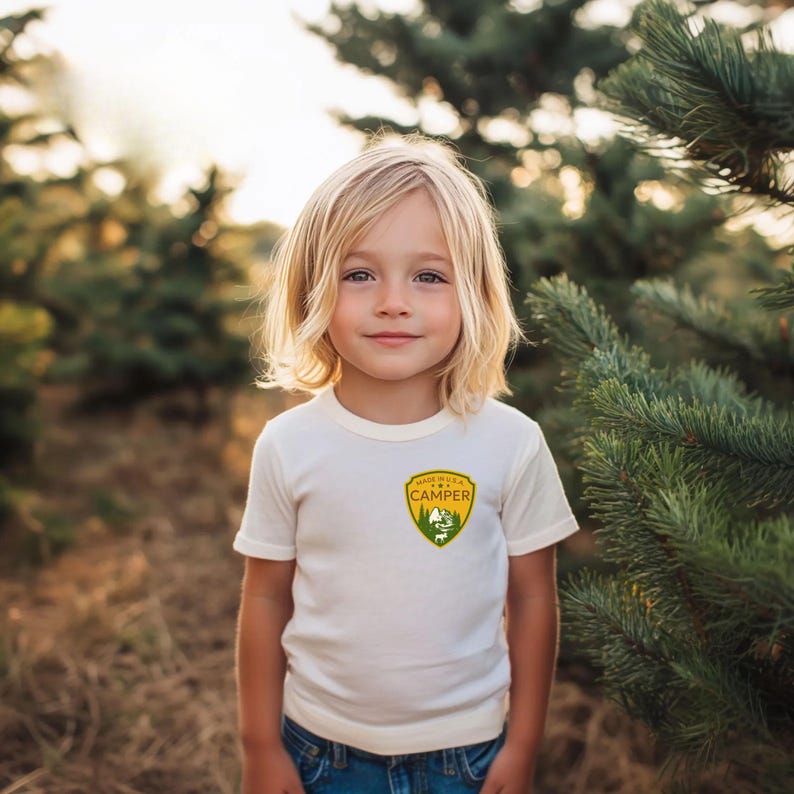 May include: A cream-colored t-shirt with a green and yellow patch that reads "Made in the USA Camper" with a mountain and tree graphic. The shirt is worn by a child with blonde hair and blue jeans.