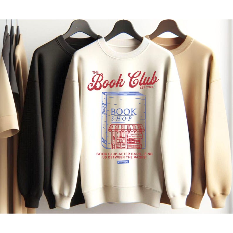 K&ouml;nnte beinhalten: Cremefarbenes Sweatshirt mit dem Text "The Book Club" in roter Schrift &uuml;ber einer Buchladen-Grafik. Die Grafik enth&auml;lt die W&ouml;rter "BOOK SHOP" und den Satz "Book Club After Dark... Find Us Between the Pages!" in einem blau-roten Design.