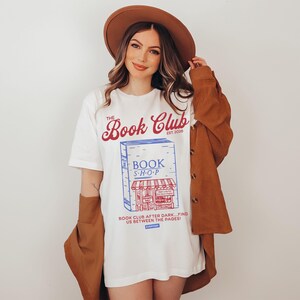 K&ouml;nnte beinhalten: Wei&szlig;es T-Shirt mit einer Grafik eines Buchladens und dem Text "The Book Club Est. 2026". Das Shirt hat auch den Text "Book Club After Dark... Find Us Between The Pages!". Die Person tr&auml;gt einen braunen Hut und eine Jacke.