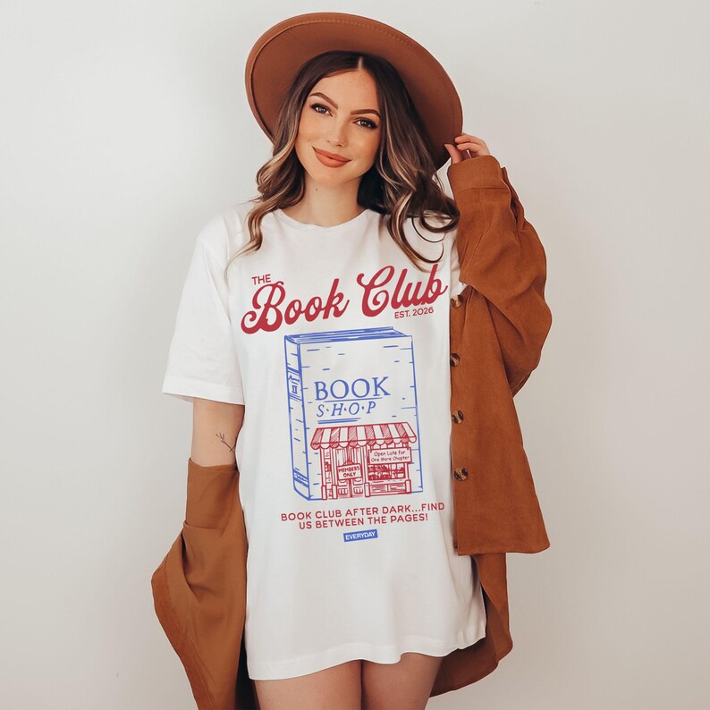 K&ouml;nnte beinhalten: Wei&szlig;es T-Shirt mit einer Grafik eines Buchladens und dem Text "The Book Club Est. 2026". Das Shirt hat auch den Text "Book Club After Dark... Find Us Between The Pages!". Die Person tr&auml;gt einen braunen Hut und eine Jacke.