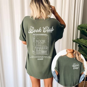 K&ouml;nnte beinhalten: Olivgr&uuml;nes T-Shirt mit wei&szlig;er Grafik eines Buchladens und dem Text "The Book Club EST. 2020." Auf der R&uuml;ckseite des Shirts steht der Text "WHAT HAPPENS BETWEEN THE PAGES STAYS BETWEEN THE PAGES!"