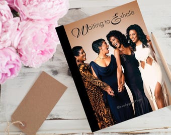 May include: A notebook with the title "Waiting to Exhale" and an image of four women in elegant dresses. The cover is on a white surface with pink flowers and a brown tag. A pencil rests on the right side of the notebook.