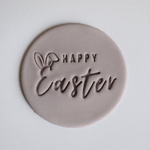 以下が含まれることがあります： 「Happy Easter」の文字とウサギの耳のデザインが施された、薄い紫色の丸いスタンプです。