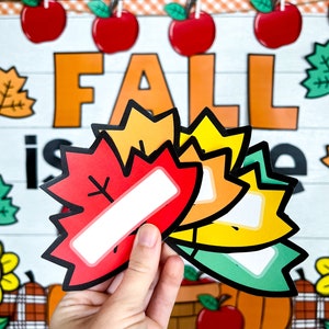 May include: Four colorful fall leaf-shaped name tags with blank spaces for writing. The leaves are red, orange, yellow, and green. The leaves are arranged in a stack with the red leaf on top. The leaves are held in a hand against a background of a fall-themed bulletin board with the word "FALL" in large orange letters.