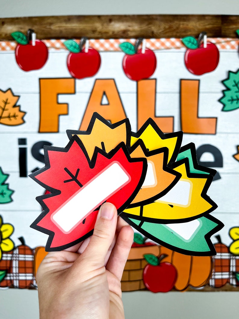 May include: Four colorful fall leaf-shaped name tags with blank spaces for writing. The leaves are red, orange, yellow, and green. The leaves are arranged in a stack with the red leaf on top. The leaves are held in a hand against a background of a fall-themed bulletin board with the word "FALL" in large orange letters.