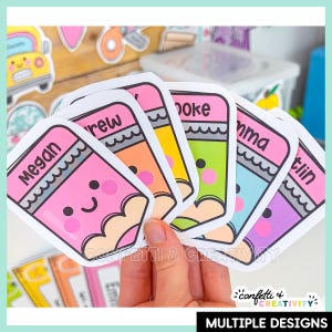 May include: A set of die-cut stickers shaped like cartoon pencils with smiling faces, each featuring a different pastel color and a personalized name printed in black text. The stickers are arranged in a fan shape, displaying various colors.