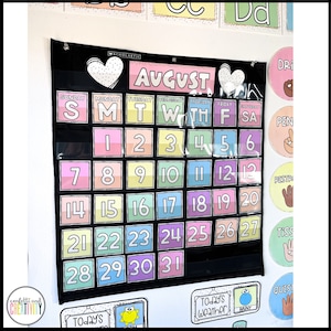 May include: A black calendar with a white heart and the word "AUGUST" in pink letters. The calendar has pockets for each day of the month, with colourful numbers in each pocket. The calendar is hanging on a white wall.