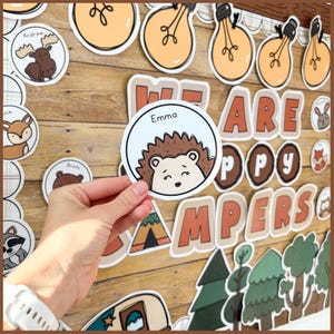 Op de afbeelding: Een ronde sticker met een illustratie van een egel en de naam "Emma" wordt voor een houten achtergrond gehouden. De achtergrond bevat decoraties met een campingthema, waaronder gloeilampen, bomen en de woorden "WE ARE HAPPY CAMPERS".