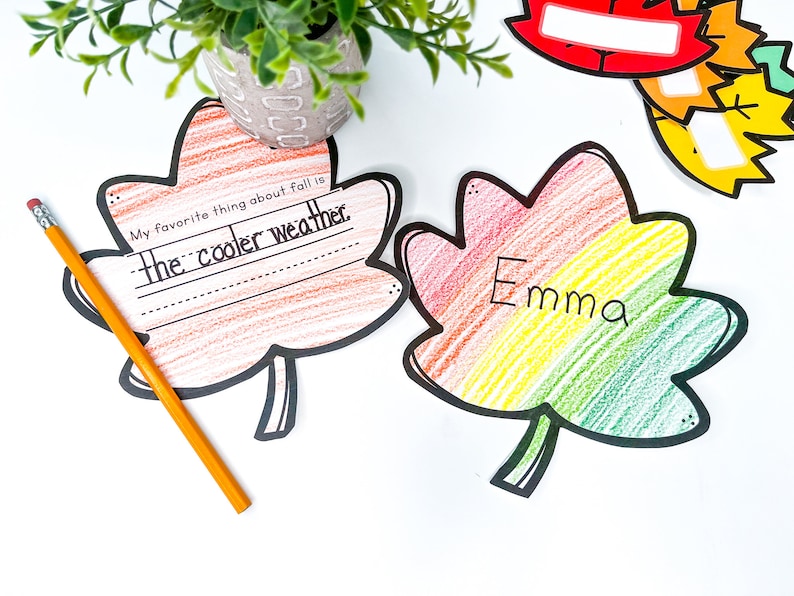 May include: Two fall-themed leaf-shaped paper crafts. One is orange and red with the text "My favorite thing about fall is the cooler weather." The other is rainbow colored with the name "Emma" written on it.