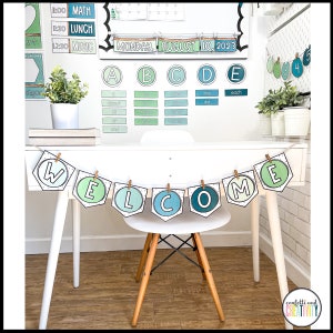 May include: A white desk with a welcome banner hanging above it. The banner is made of green, blue, and white paper and has the word "WELCOME" written on it. The desk is decorated with a calendar, alphabet chart, and other classroom decorations. There is a chair in front of the desk.