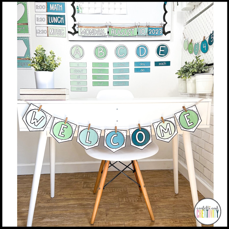 May include: A white desk with a welcome banner hanging above it. The banner is made of green, blue, and white paper and has the word "WELCOME" written on it. The desk is decorated with a calendar, alphabet chart, and other classroom decorations. There is a chair in front of the desk.