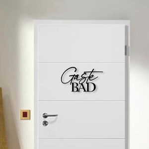 May include: A white door with a black sign that says "G&auml;ste Bad" which translates to "Guest Bathroom" in English. The door has a silver door handle and a small red light switch on the wall next to it. There is a white couch with a white throw blanket and a painting of a yellow rose on the wall behind it.