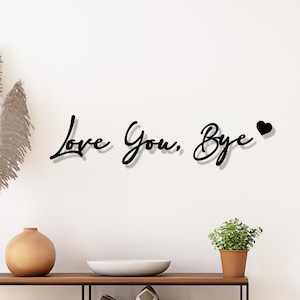 Könnte beinhalten: Schwarze Metall-Wandkunst mit der Aufschrift "Love You, Bye" und einem Herzsymbol. Der Text ist in einer Schreibschrift gehalten. Die Wandkunst ist an einer weißen Wand über einem Holzregal angebracht.