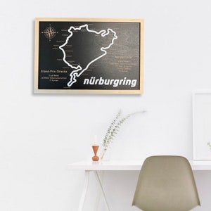 May include: A wooden sign with a black background and a white outline of the N&uuml;rburgring race track. The sign also includes the text "Nordschleife", "Grand-Prix-Strecke", "n&uuml;rburgring", and other details about the track.