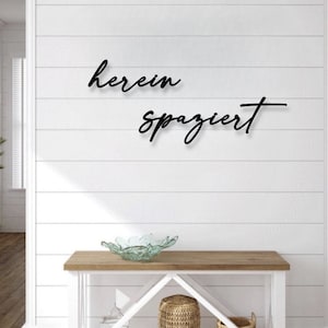 Könnte beinhalten: Schwarze Wanddekoration aus Metall mit den Worten "herein spaziert" in einer kursiv geschriebenen Schrift. Die Dekoration ist an einer weißen Schiffslappenwand über einem Holztisch mit einer Glasschale und einem geflochtenen Korb angebracht.