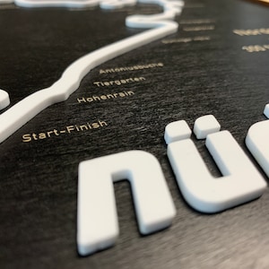 May include: A black wooden sign with a white outline of a race track. The sign says "N&uuml;r" in large white letters and includes the text "Start-Finish", "Hohenrain", "Tiergarten", and "Antoniusbuche".