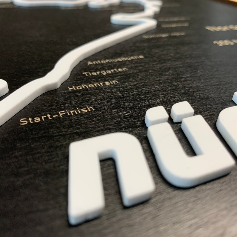 May include: A black wooden sign with a white outline of a race track. The sign says "N&uuml;r" in large white letters and includes the text "Start-Finish", "Hohenrain", "Tiergarten", and "Antoniusbuche".