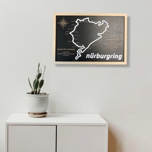 May include: A black and white wooden sign with a map of the N&uuml;rburgring race track. The sign is framed in a light wood frame and has the text "N&uuml;rburgring" in white letters.