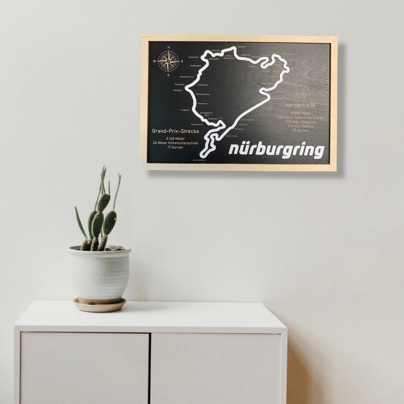 May include: A black and white wooden sign with a map of the N&uuml;rburgring race track. The sign is framed in a light wood frame and has the text "N&uuml;rburgring" in white letters.
