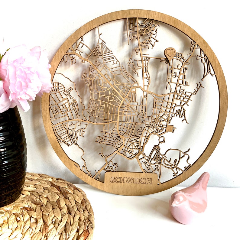 May include: A wooden map of Schwerin, Germany, with a heart-shaped marker in the center. The map is circular and has a light brown color.
