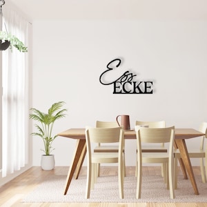 May include: Black metal wall decor featuring the text "Ess Ecke" in a contemporary, cursive font. The decor is mounted on a white wall above a wooden dining table with four chairs.
