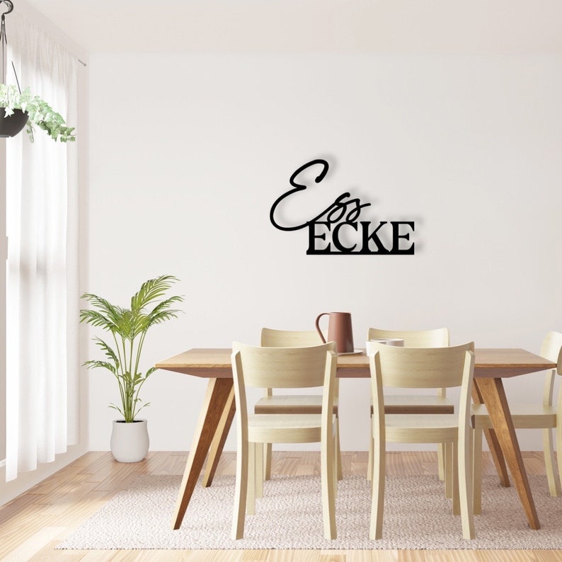 May include: Black metal wall decor featuring the text "Ess Ecke" in a contemporary, cursive font. The decor is mounted on a white wall above a wooden dining table with four chairs.