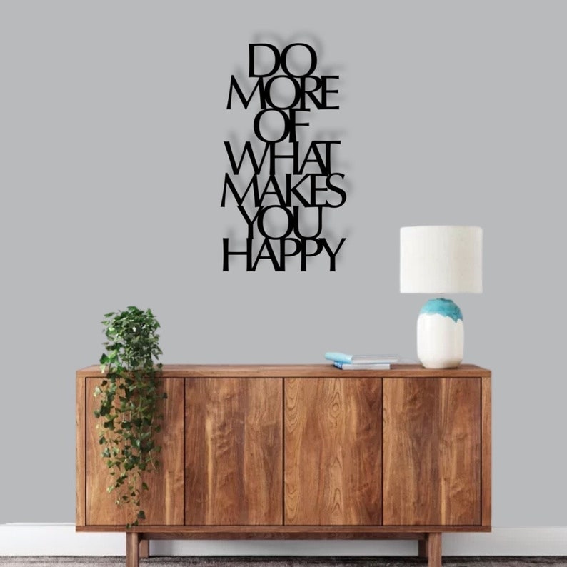 May include: Black metal wall art with the text "Do More Of What Makes You Happy". The art is hanging on a gray wall above a wooden dresser with a lamp on the right side.