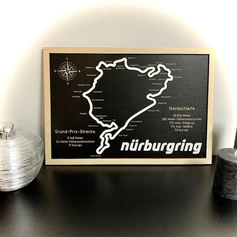 May include: A black wooden sign with a white outline of the N&uuml;rburgring race track. The sign includes the names "Grand-Prix-Strecke" and "Nordschleife" with details about the track's length, elevation change, and number of turns.
