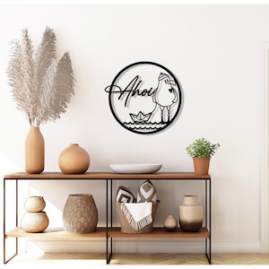 Könnte beinhalten: Schwarze Metall-Wandkunst mit einer Möwe und einem Papierboot in einem Kreis, mit dem Wort "Ahoi" in Schreibschrift. Das Kunstwerk ist an einer weißen Wand über einer hölzernen Konsole angebracht.