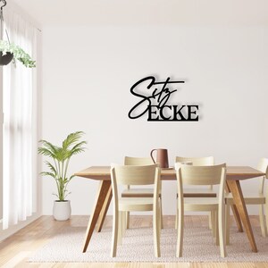 May include: Black metal wall decor featuring the text "City Ecke" in a contemporary script font. The decor is mounted on a white wall above a wooden dining table with four chairs.