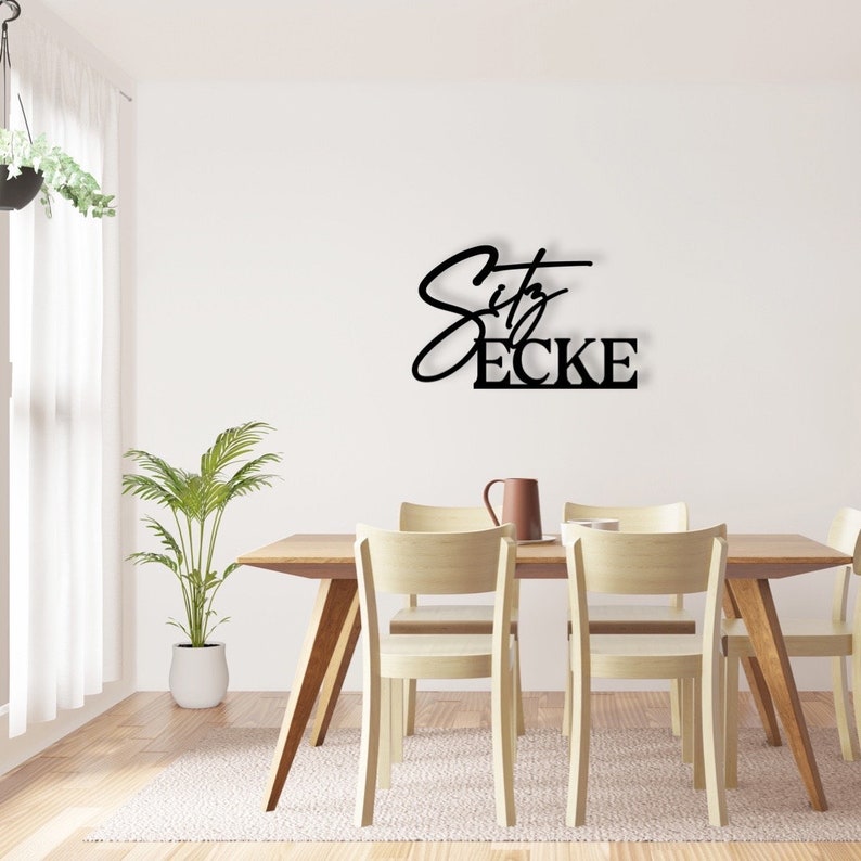 May include: Black metal wall decor featuring the text "City Ecke" in a contemporary script font. The decor is mounted on a white wall above a wooden dining table with four chairs.