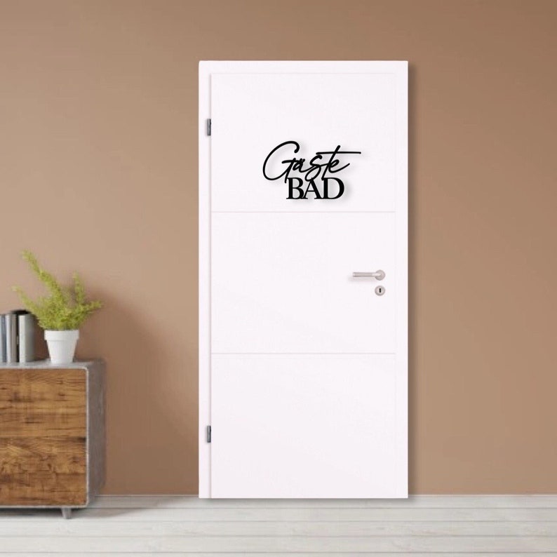 May include: White door with a black sign that says "G&auml;ste Bad" which translates to "Guest Bathroom" in English.