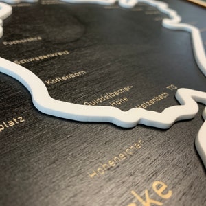 May include: A black wooden map with white raised lines outlining the shape of a region. The map features various place names in gold lettering, including "Kottenborn", "Quiddelbacher Hohe", and "Hatzbach Trg".