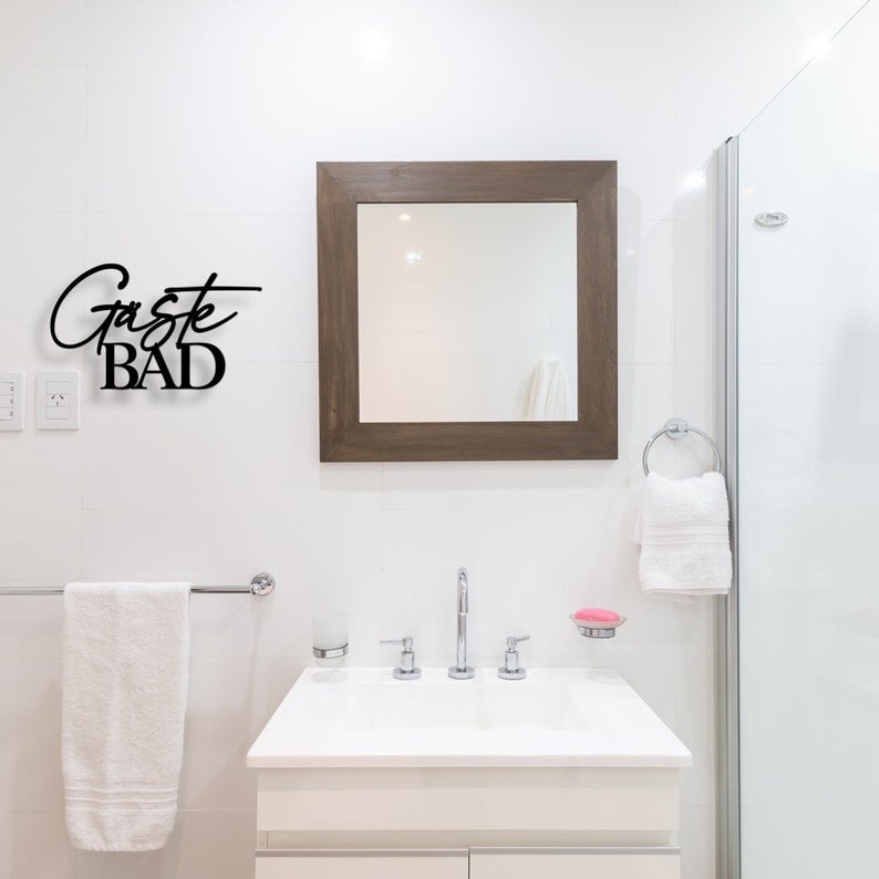 May include: A white bathroom with a white towel hanging on a towel rack, a white sink with a chrome faucet, a mirror with a brown frame, and a sign that says "G&auml;ste BAD" in black lettering.